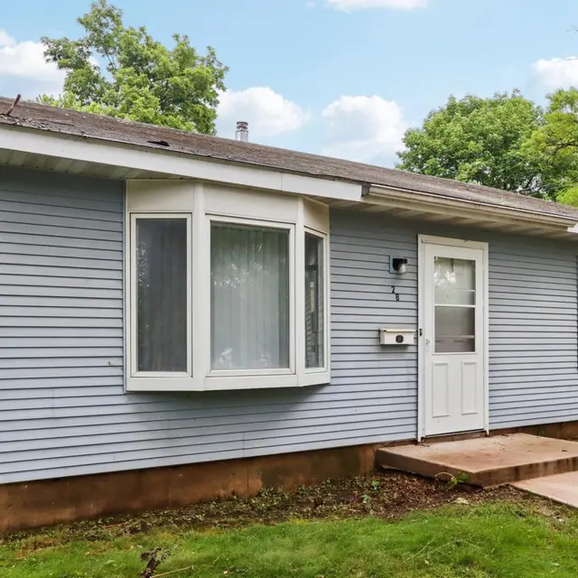 A small blue house with a flat roof, featuring a large bay window and a front door. The house is surrounded by green grass and trees in the background.