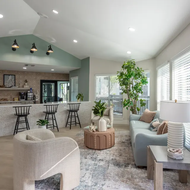 A modern living space featuring a cozy seating area with a light blue sofa and a round coffee table. In the background is a kitchen bar with high chairs, decorated with plants. Natural light pours in through large windows, enhancing the airy atmosphere.