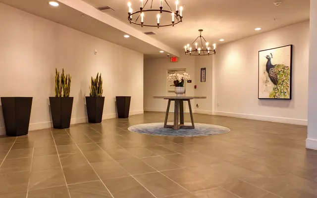 A modern lobby interior with a round table at the center, surrounded by tall plants in black pots. There are two stylish light fixtures hanging from the ceiling, and a decorative artwork featuring a peacock on the wall.