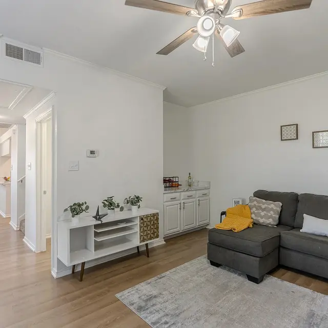 A cozy living room featuring a gray sectional sofa with decorative pillows, a light gray area rug, and a white cabinet with plants. The wall has framed pictures. In the background, there's a hallway leading to a dining area with windows.