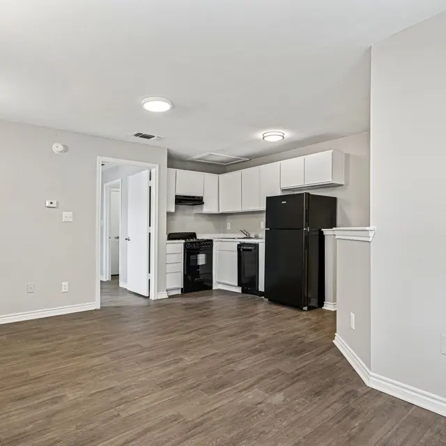 A contemporary kitchen and living space with a light-colored wall and wooden flooring. The kitchen features white cabinetry and black appliances, including a refrigerator and oven. A doorway leads to another room, and ample natural light comes from the window.