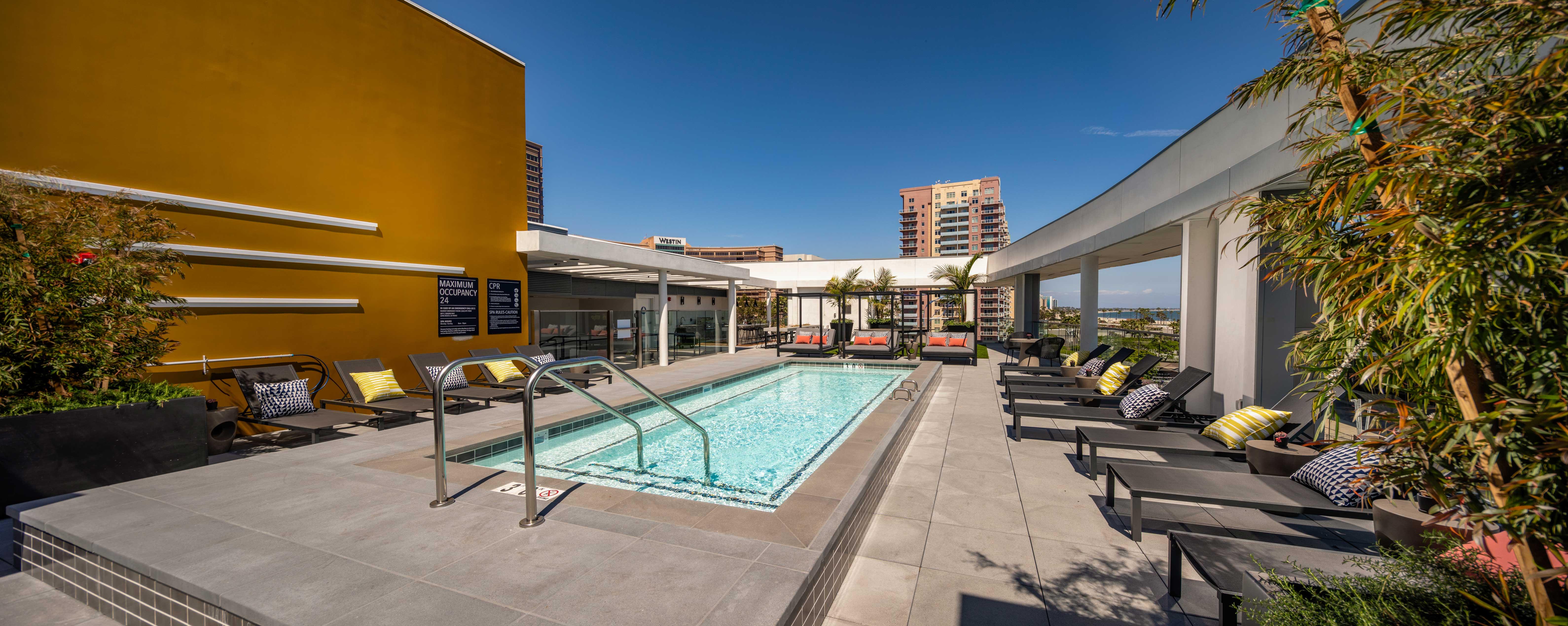 A modern rooftop pool area with a clear swimming pool, lounge chairs, and vibrant yellow wall against a clear blue sky.