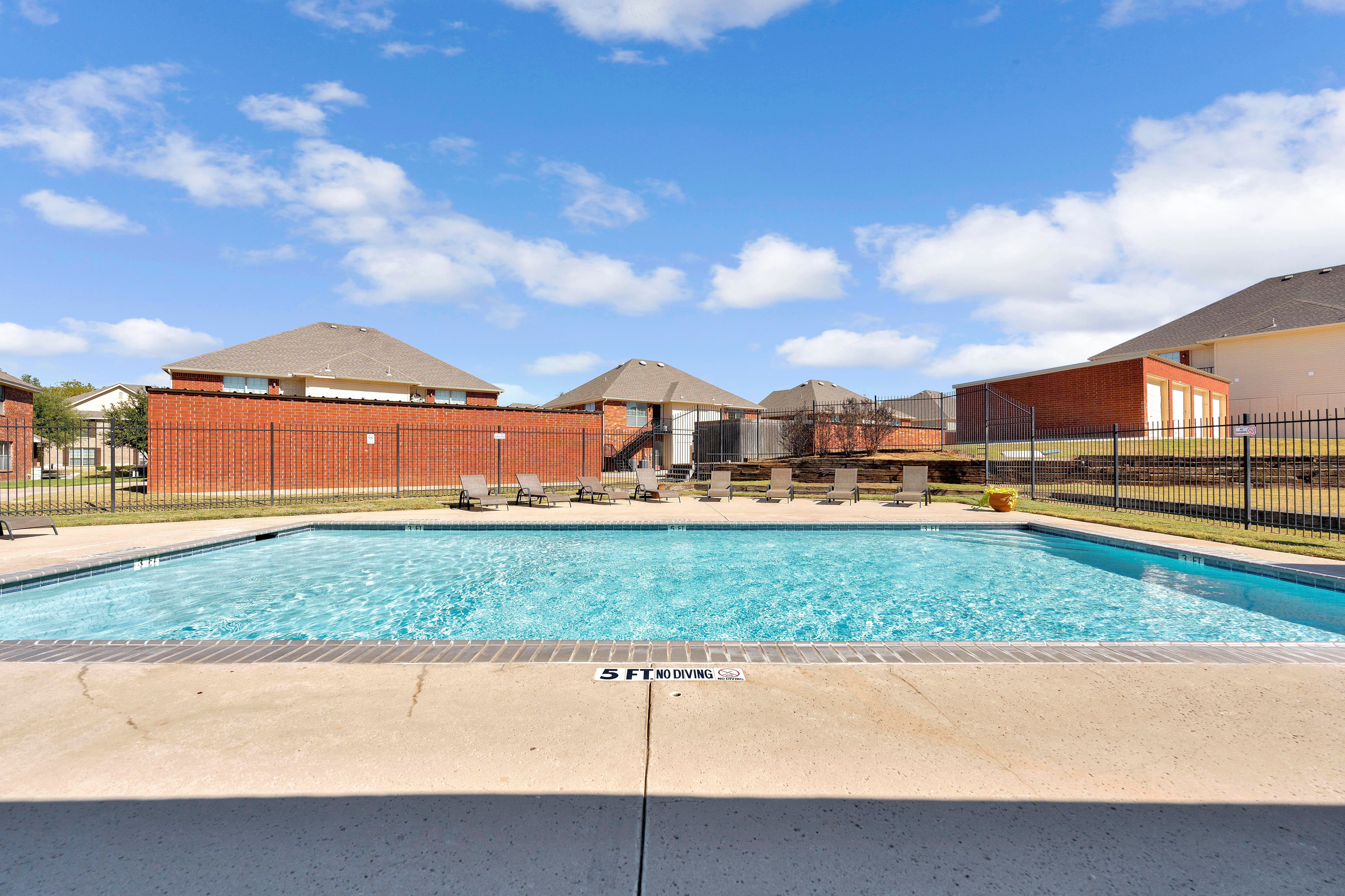 A swimming pool in a residential area, surrounded by a fence with lounge chairs and buildings in the background under a clear blue sky.