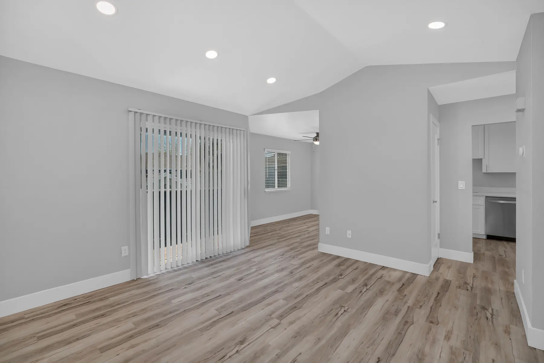 A modern living room featuring light gray walls, large windows with vertical blinds, and light wood flooring. The space is bright with recessed lighting and an open layout leading to a kitchen area on the right.