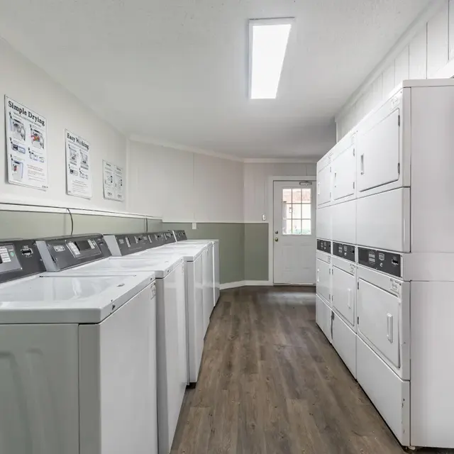 Interior of a laundry room featuring multiple washing machines along a wall, with a door leading outside and storage cabinets on the opposite wall.