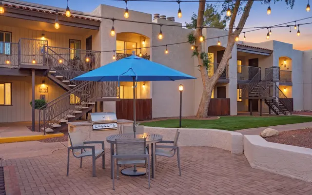 Outdoor lounge area featuring a table with chairs and a blue umbrella, surrounded by string lights and an apartment complex in the background.