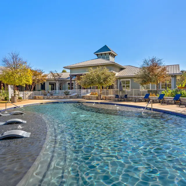 A luxurious swimming pool surrounded by lounge chairs and greenery, with a clubhouse in the background under a clear blue sky.