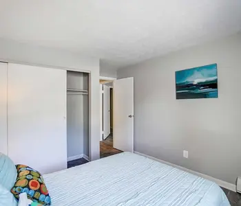 A neatly arranged bedroom featuring a bed with light blue bedding, a colorful pillow, and a bedside lamp. There's a closet with sliding doors on the left and a door leading to another room on the right. A piece of wall art is displayed above the bed.