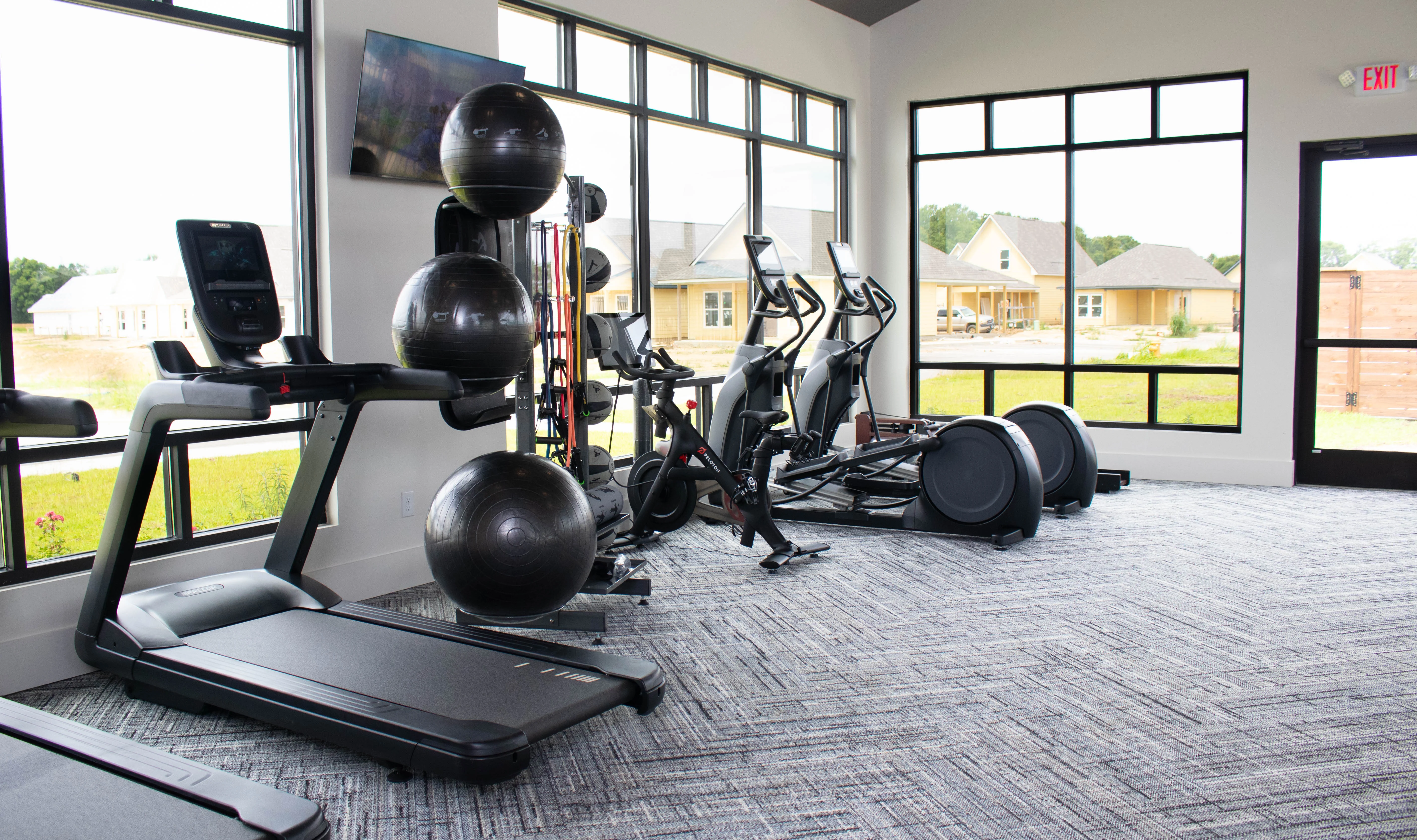 A modern fitness room with various gym equipment, including a treadmill, elliptical trainers, exercise balls, and a large window with a view outside.