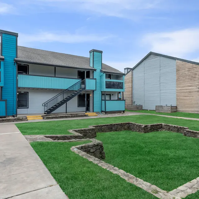 A view of a residential apartment complex with two buildings, featuring blue accents and a well-maintained lawn. There are walkways and staircases leading to the apartments.