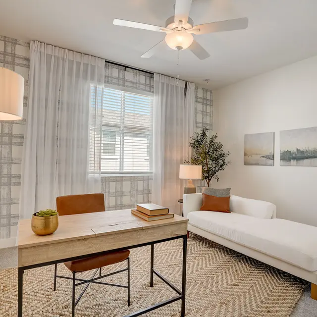 Cozy Modern Home Office A modern home office featuring a beige rug, white chaise lounge, wooden desk with a plant and books, and a large window with sheer curtains.