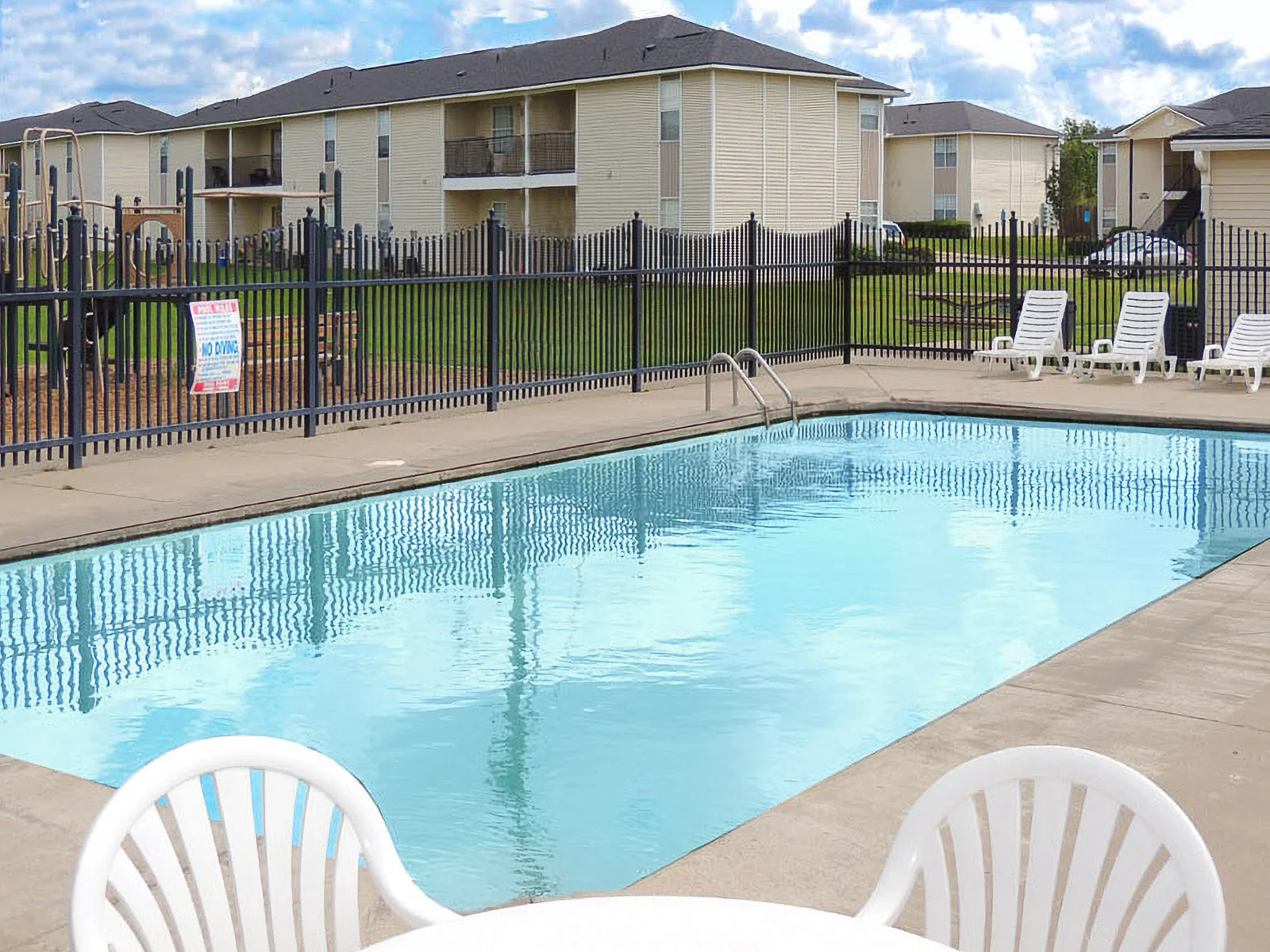 Community Pool with Apartment Views An outdoor swimming pool with clear water surrounded by a fence, lounge chairs, and apartment buildings in the background.