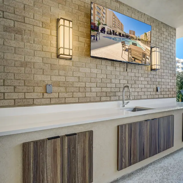 A stylish outdoor kitchen area with a minimalist design featuring a large countertop, a sink, and wooden cabinet doors. A flat-screen TV is mounted on the brick wall with a view of a pool area in the background.