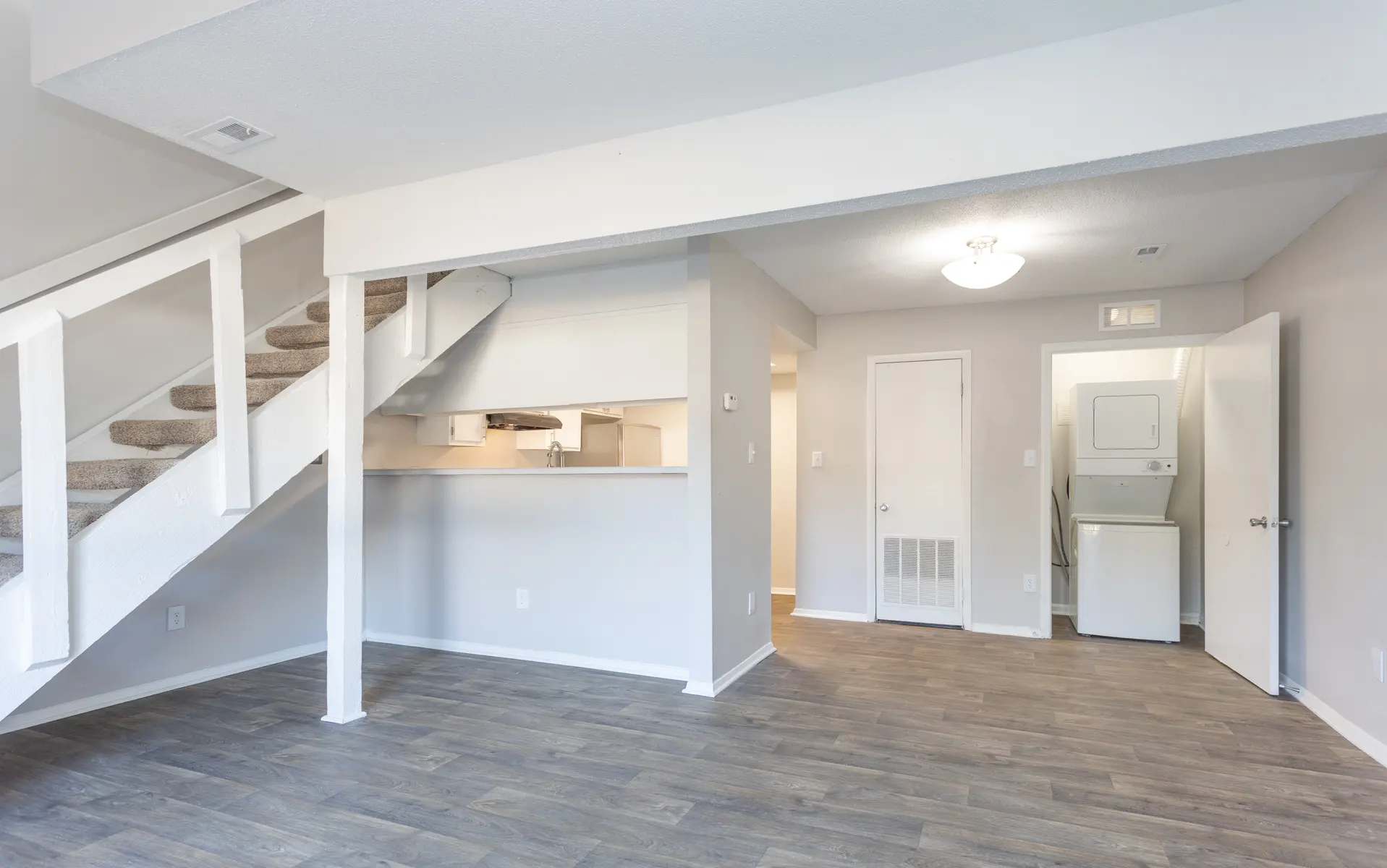 A spacious living area featuring a staircase, light-colored walls, and wooden flooring. The scene shows an open floor plan with a view of a kitchen area and laundry room.