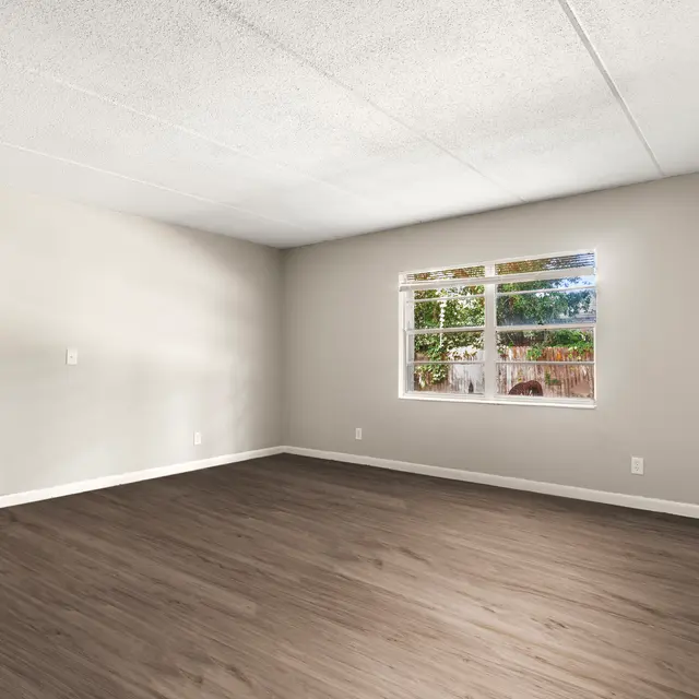 An empty room with light gray walls, a window with a garden view, and a door on the left side.