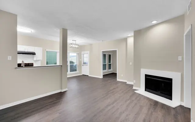 A spacious living room with light-colored walls and dark wooden floors. A fireplace is visible on the right side. There is a small dining area with a chandelier in the back, and a kitchen area with white cabinets in the background. Large windows allow natural light to enter the room.