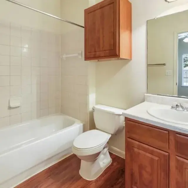A clean and modern bathroom featuring a white bathtub, a toilet, and a sink with a wooden cabinet. The walls are tiled, and there is a mirror above the sink. A window shows greenery outside.