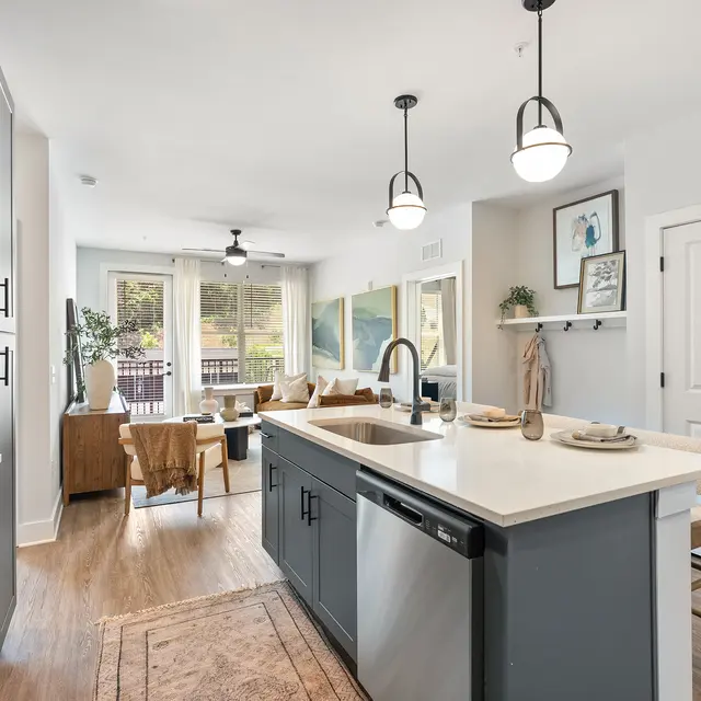A modern kitchen featuring an island with a sink, gray cabinets, and stainless steel appliances. The space is bright with large windows and wooden flooring, connecting to a living area with comfortable seating.