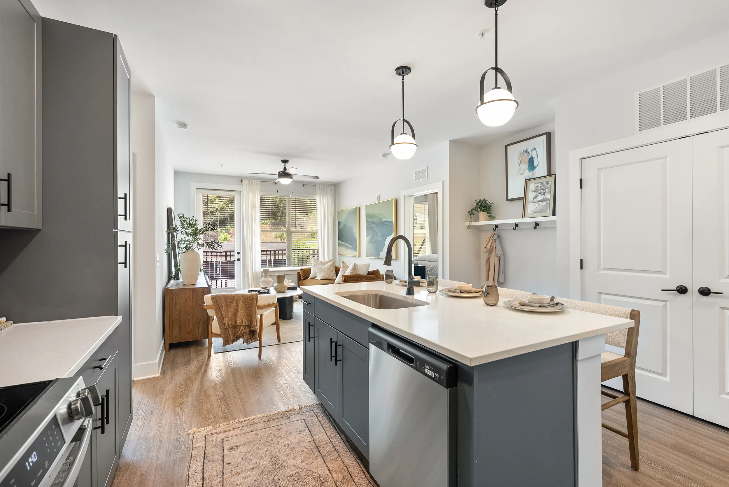 Contemporary Kitchen Interior A modern kitchen featuring an island with a sink, gray cabinets, and stainless steel appliances. The space is bright with large windows and wooden flooring, connecting to a living area with comfortable seating.