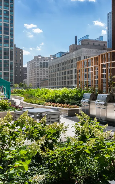 A spacious rooftop garden with several stainless steel grills, surrounded by greenery and planted beds. Tall buildings are visible in the background under a blue sky with scattered clouds.
