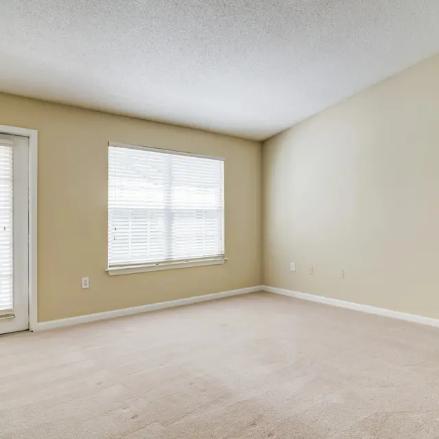 An empty room featuring beige walls and light-colored carpet. It has a door on the left side leading outside, and a window with white blinds situated in the middle, letting natural light in.