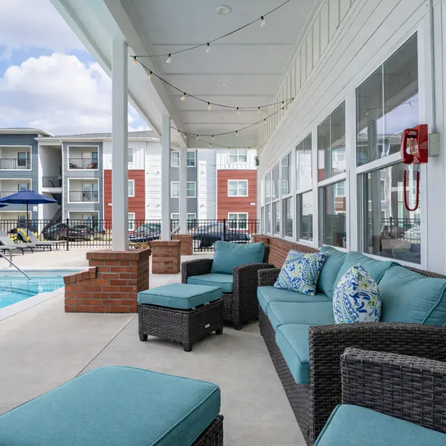 A stylish poolside lounge area with teal cushions, wicker furniture, and a swimming pool in the background framed by apartment buildings.
