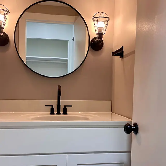 A modern bathroom featuring a circular mirror above a white sink with black fixtures. Two vintage-style light fixtures are mounted on the wall. A partially open door leads to a closet space.
