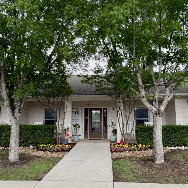 A welcoming entrance to a building surrounded by trees and flower beds. The pathway leads to double doors that are framed by greenery.