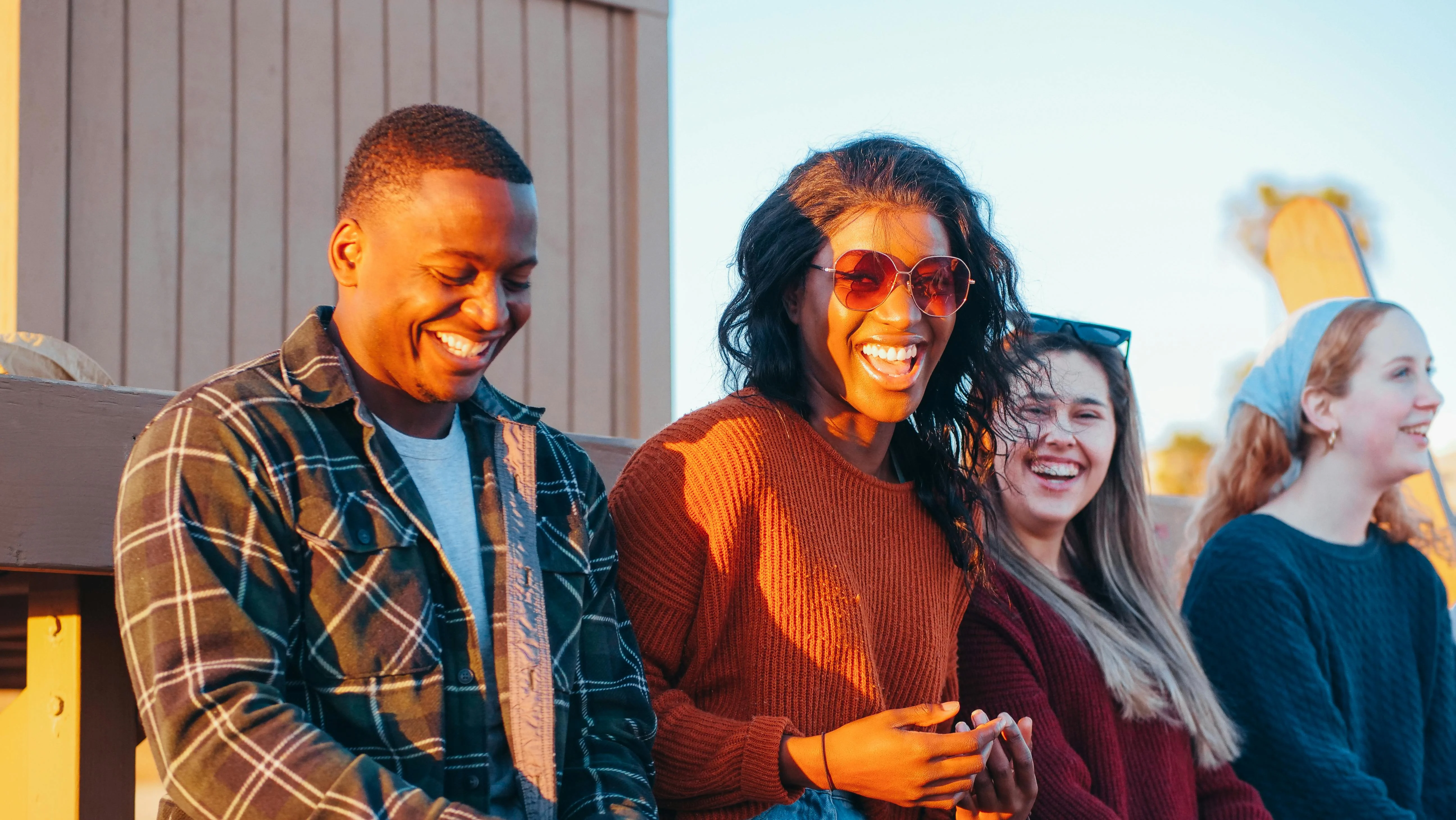 Joyful Friends Outdoors A group of four friends laughing and enjoying a sunny day outdoors. They are seated on a bench with smiles on their faces. The first person on the left is wearing a plaid jacket, the second person has sunglasses on and is in a cozy sweater, the third person has long hair and is smiling, while the last person on the far right is wearing a beanie.