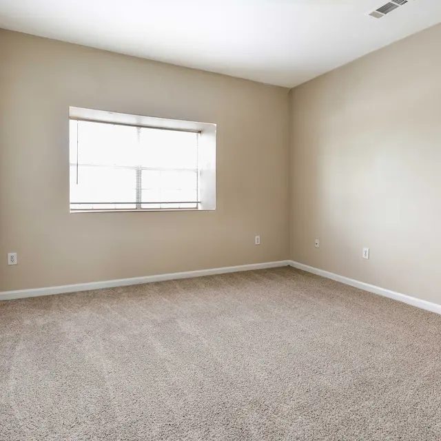 An empty room with beige walls and carpet, featuring two white doors and a window with natural light coming in.