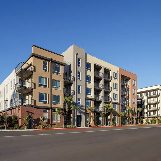 A modern multi-story apartment building with a colorful facade, situated on a sunny day with a clear blue sky. The building features balconies and palm trees in the foreground, with mountains visible in the background.