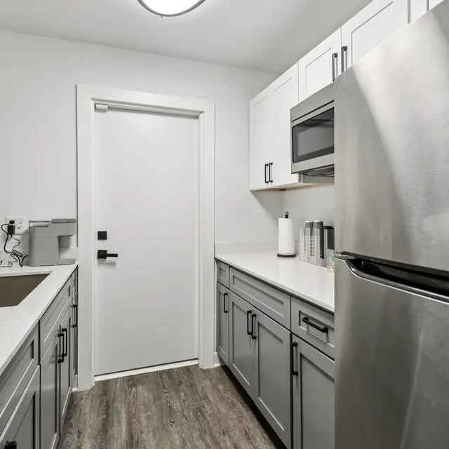 A modern kitchen featuring stainless steel appliances, gray cabinetry, and a white countertop. There's a sink with a black faucet and a door leading outside in the background.