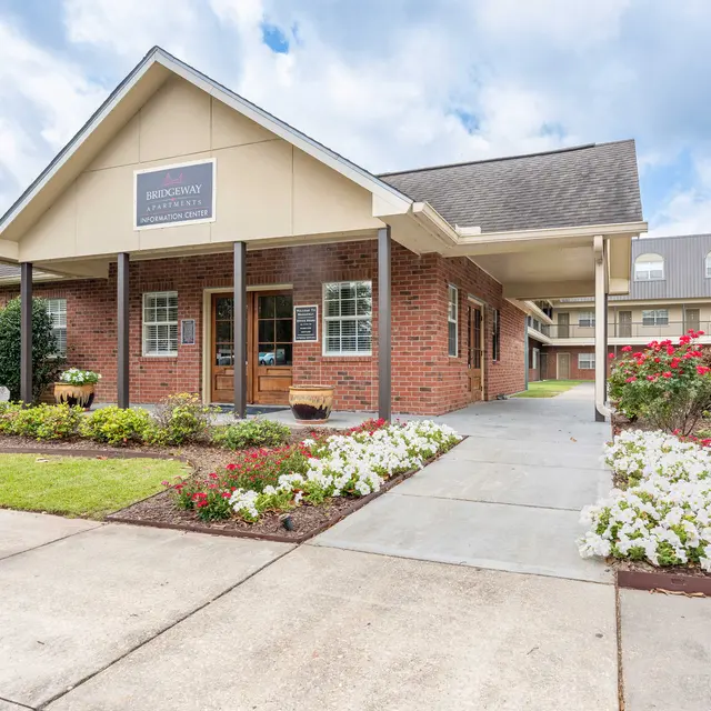 Exterior view of a brick building with a porch, surrounded by greenery and flowers, under a cloudy sky.