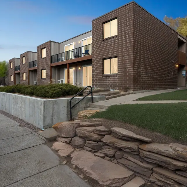Modern Apartment Complex A modern apartment building with a mix of brick and siding exteriors, featuring balconies and landscaped areas. The scene captures a corner view with a pathway leading up to the entrance.