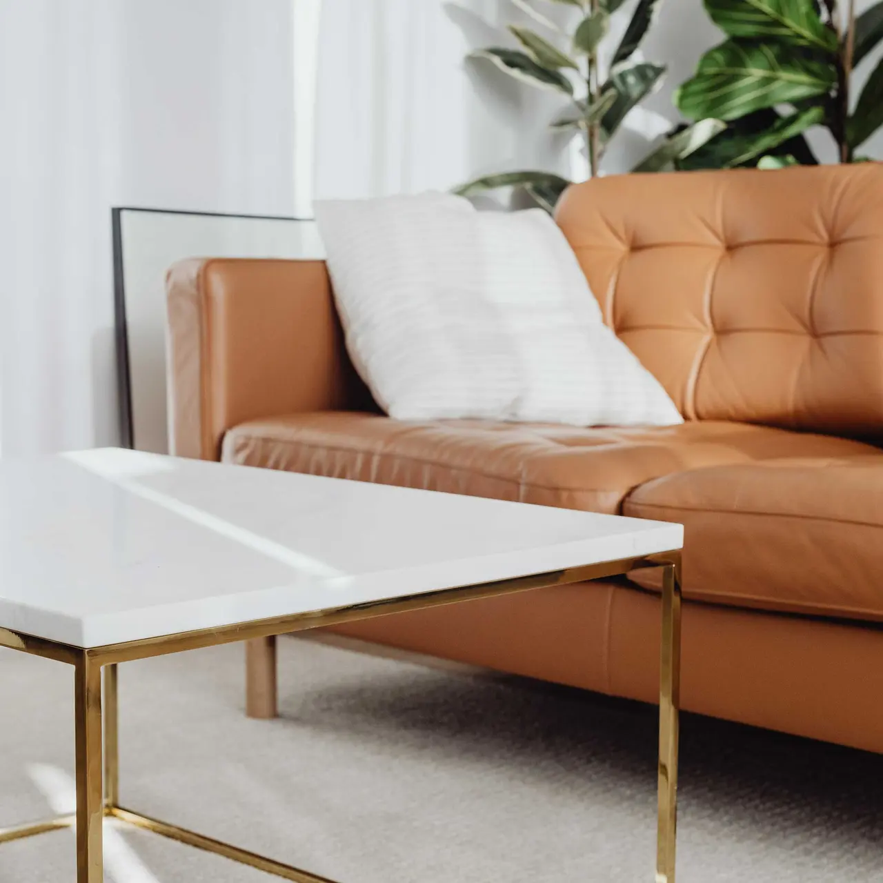 Modern Living Room Setup A brown leather sofa with cushions beside a white coffee table on a light-colored rug, framed by plants and natural light.