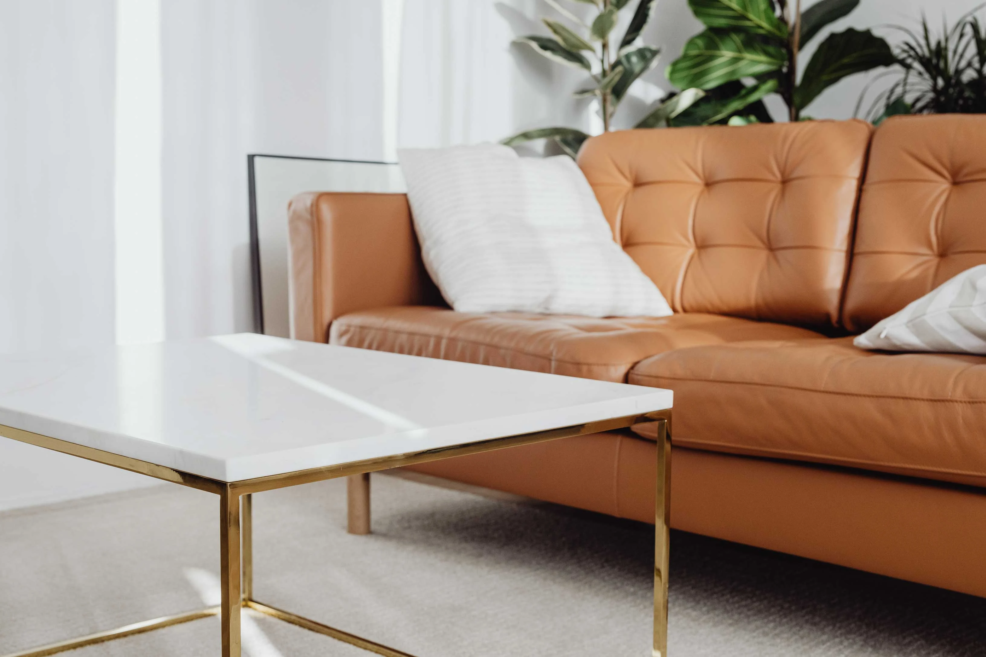 Modern Living Room Setup A brown leather sofa with cushions beside a white coffee table on a light-colored rug, framed by plants and natural light.