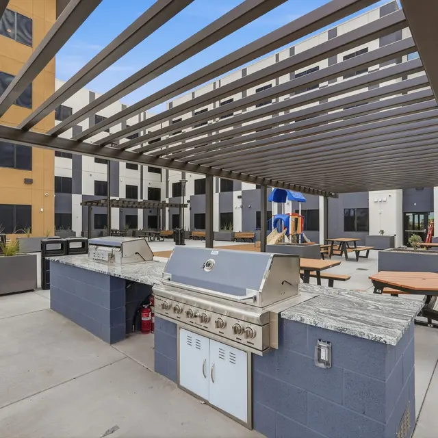 Outdoor Grill Area in Courtyard An outdoor grill area with picnic tables under a pergola, located in a courtyard of a modern building.