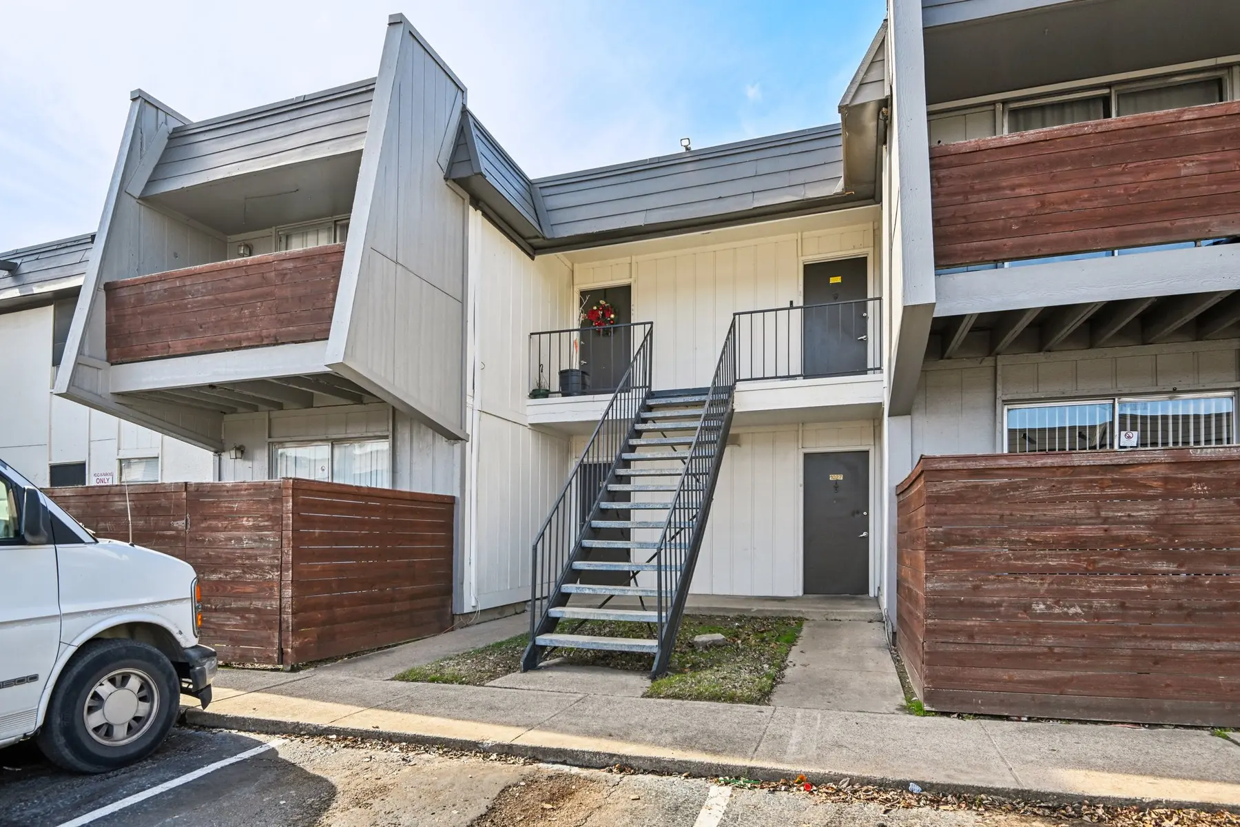 Exterior view of a two-story apartment building with stair access. Two levels with balconies and wooden fencing around the ground floor. A white van is parked in front.