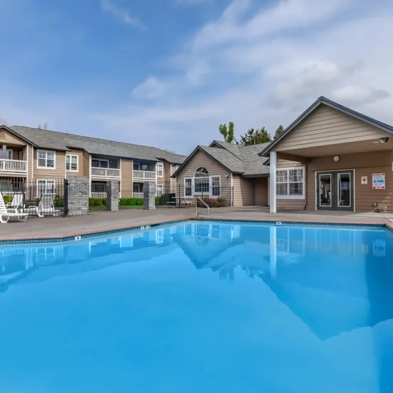 A clear, blue swimming pool in front of a residential building complex with lounge chairs and an outdoor clubhouse.