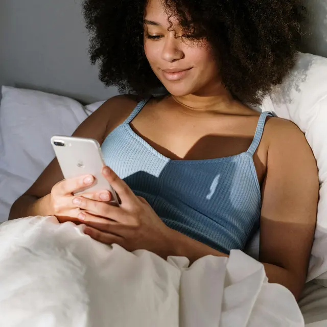 Woman Using Phone in Bed A woman with curly hair sitting in bed, holding a smartphone and looking at the screen with a relaxed smile.