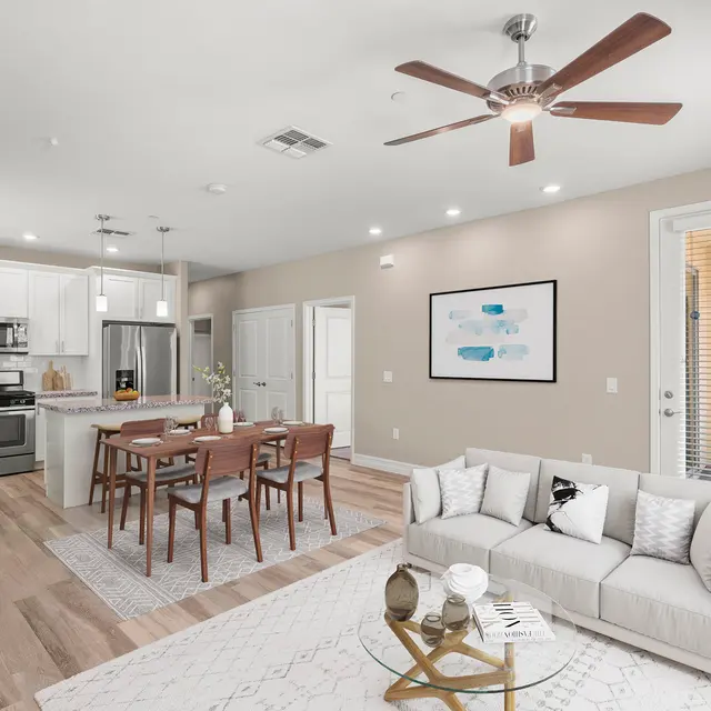 A modern open-concept living space featuring a light-colored sofa, wooden dining table, and a kitchen visible in the background.