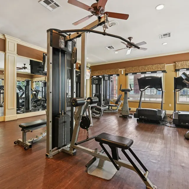 A well-lit gym interior featuring various workout machines and exercise equipment. The room has a hardwood floor, large mirrors, and multiple windows allowing natural light. There are cardio machines, strength training equipment, and fans on the ceiling.