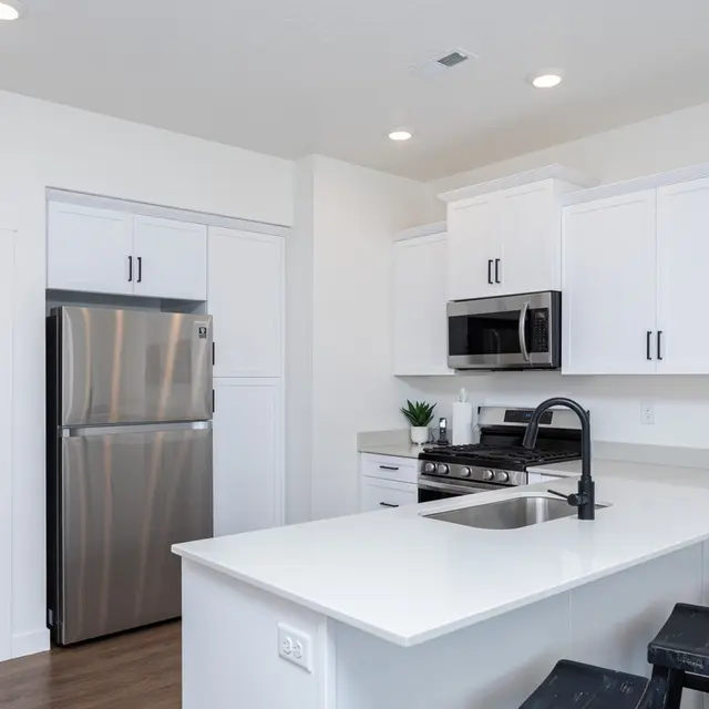 A modern kitchen featuring white cabinetry, stainless steel appliances, and a spacious countertop with a kitchen island. There are light fixtures and wooden flooring.