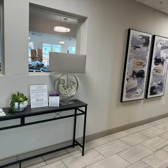 A modern hallway featuring a black console table with decorative items, two lamps, and a small plant. There are two artworks on the wall, and the space is well-lit with large windows in the background.