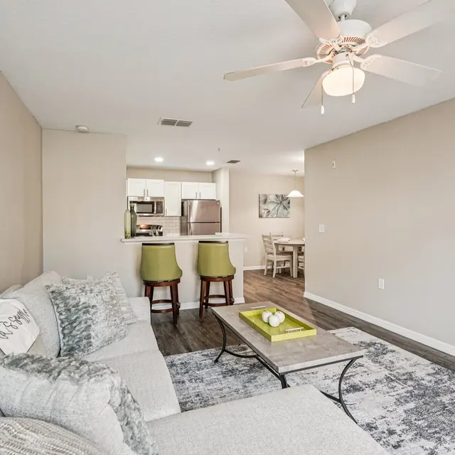 A modern living room featuring a light gray sectional sofa, a decorative coffee table, and a ceiling fan. The space leads to a kitchen area with bar stools and a dining area in the background.