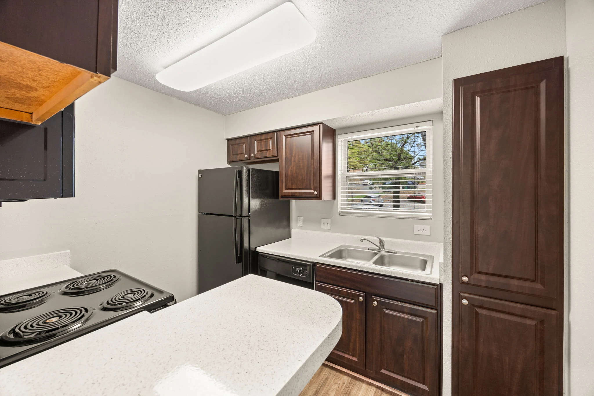 Modern Kitchen Interior A modern kitchen featuring dark wood cabinets, a black refrigerator, a white countertop, and a stove with four burners. There is a window with a view outside and a sink integrated into the countertop.