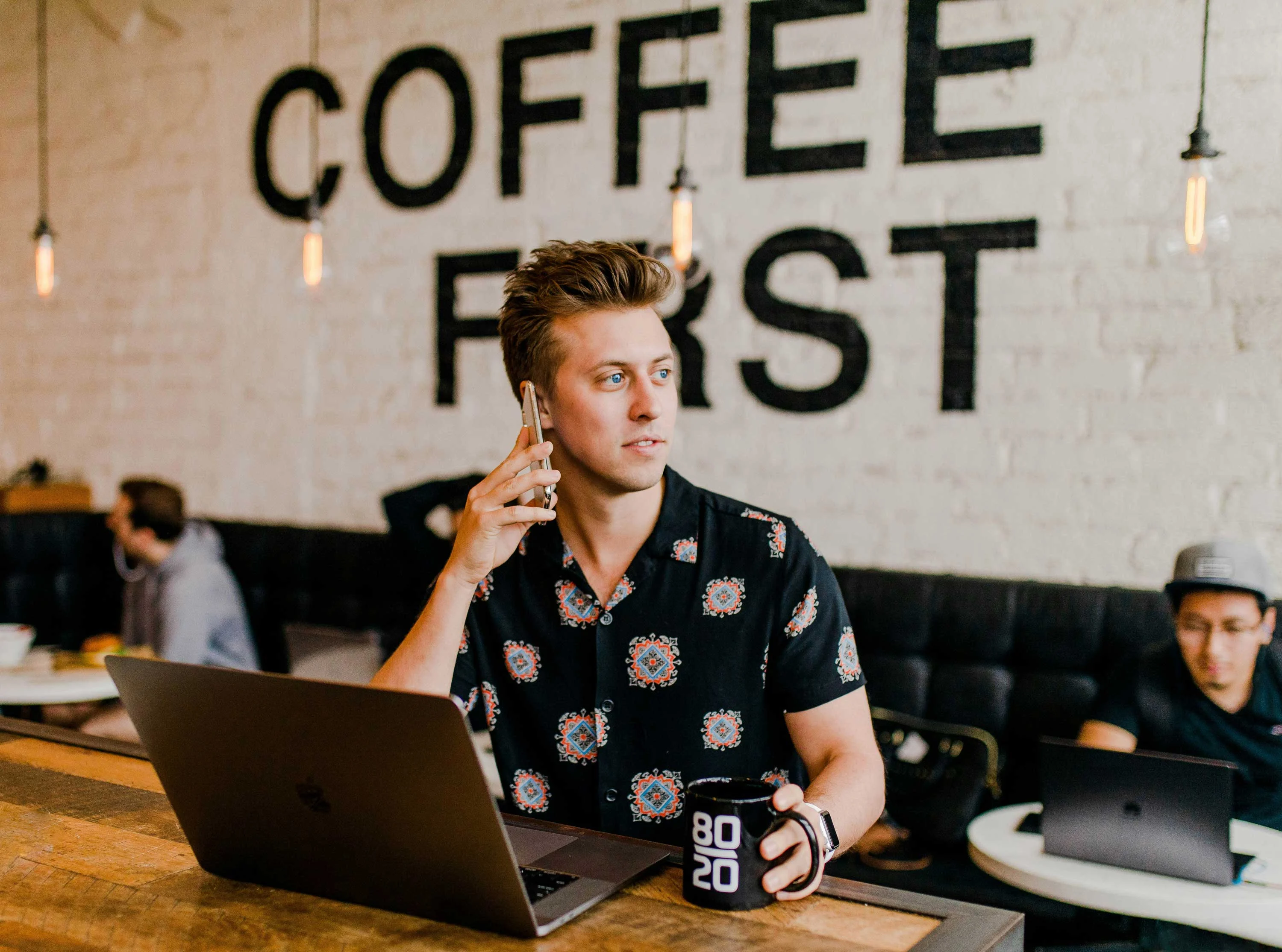 A young man on a phone call while sitting at a wooden table with a laptop and a coffee mug, in a café setting with a mural saying 'COFFEE FIRST' in the background.
