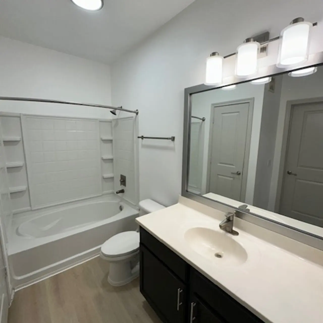 A modern bathroom featuring a bathtub with a white tiled wall, a toilet, a double vanity with a sink, and a large mirror. The room is well-lit with a ceiling light and three light fixtures above the vanity.
