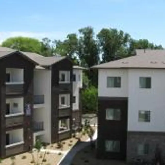 Exterior view of a modern apartment complex featuring three-story buildings with a combination of white and brown sidings, a landscaped path, and greenery in the background.