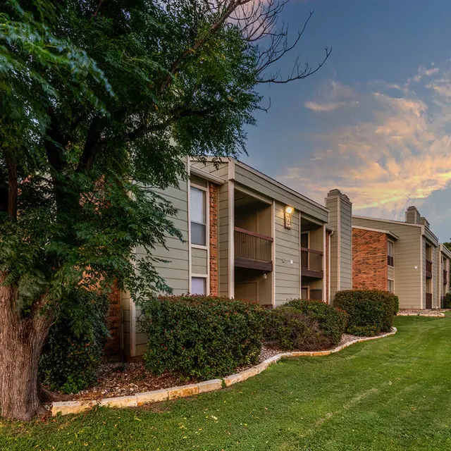 Exterior view of a multi-unit apartment complex surrounded by well-maintained landscaping and grass, with a scenic sky at dusk.